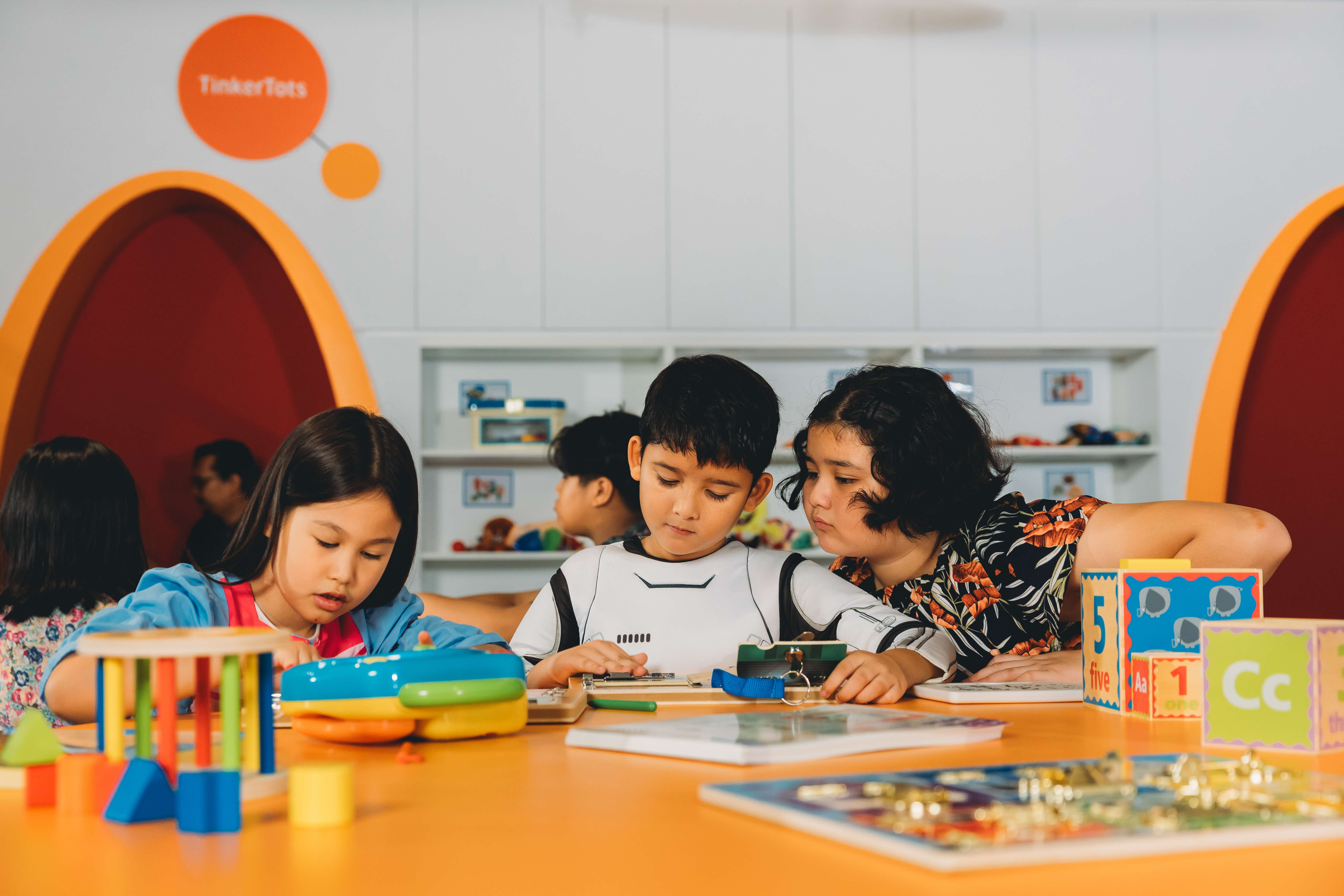 A girl and boy are seated at a table in TinkerTots focused on their building activity while another girl looks on curiously.